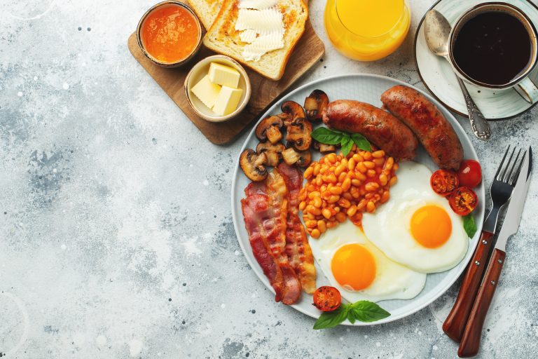 Full English breakfast on a plate with fried eggs, sausages, bacon, beans, toasts and coffee on light stone background. With copy space. Top view.